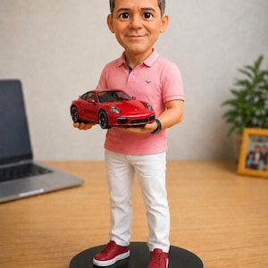 Könnte beinhalten: Eine personalisierte Wackelkopf-Figur einer Person mit kurzem braunem Haar, die ein hellrosa Poloshirt, eine weiße Hose und rote Sneaker trägt. Die Figur steht auf einem runden schwarzen Sockel und hält ein kleines rotes Sportwagenmodell in den Händen.