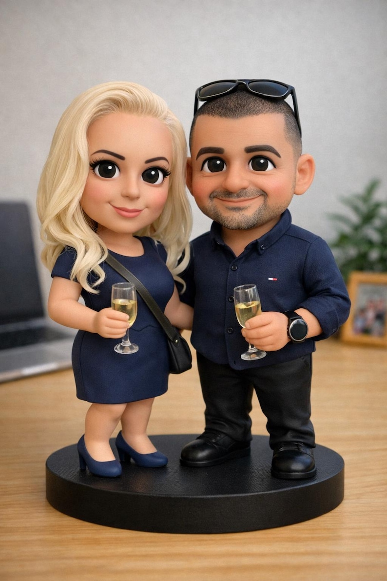 May include: A figurine of a couple, both holding champagne glasses. The woman wears a navy blue dress and heels, and the man wears a navy blue shirt and black pants. They stand on a black circular base. Sunglasses rest on the man's head.