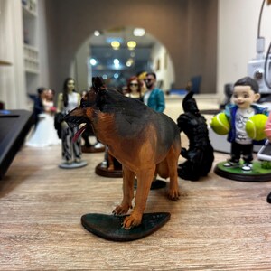 May include: A detailed figurine of a German Shepherd dog, with brown and black fur, standing on a dark green base. Other figurines of people are visible in the background, suggesting a collection of miniature figures.