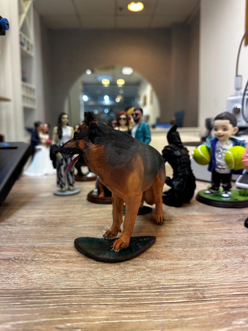 May include: A detailed figurine of a German Shepherd dog, with brown and black fur, standing on a dark green base. Other figurines of people are visible in the background, suggesting a collection of miniature figures.