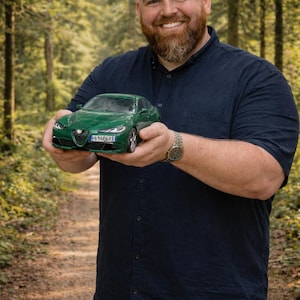 May include: A green model car with the license plate "E6486KE" is held in front of a blurred forest background. The car is a detailed replica, showcasing headlights, wheels, and a sleek design. The image is well-lit, highlighting the car's features.