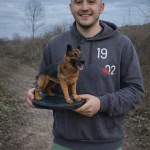 May include: A detailed sculpture of a German Shepherd dog, featuring black and tan fur, is displayed on a round, dark-colored base. The dog's tongue is visible. The sculpture is held by a person wearing a gray hoodie with the numbers "19 22" printed on it.