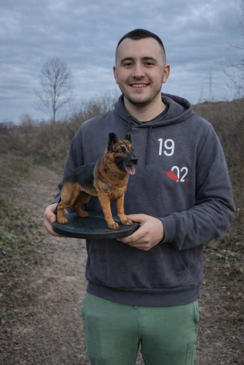 May include: A detailed sculpture of a German Shepherd dog, featuring black and tan fur, is displayed on a round, dark-colored base. The dog's tongue is visible. The sculpture is held by a person wearing a gray hoodie with the numbers "19 22" printed on it.
