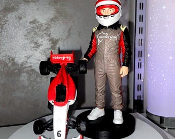 May include: A 3D printed figurine of a race car driver wearing a white helmet with red accents and a grey racing suit with black and red sleeves. The driver stands next to a red and white race car featuring the number 6 and the text nürburgring.