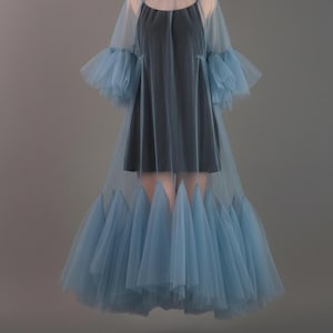 May include: A layered dress featuring a black slip dress under a sheer, light blue tulle overlay. The overlay has a high ruffled collar, bell sleeves, and a flared skirt with pointed edges. The dress is displayed on a mannequin against a gray backdrop.