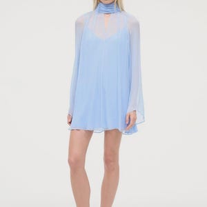 May include: A pale blue, long-sleeved dress featuring a high neckline with a keyhole. The dress has a flowing, layered design and a short hemline. The model is wearing sandals with a snakeskin pattern.