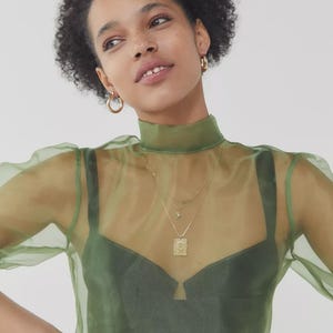 Pode incluir: Uma mulher usando um top transparente verde-oliva com gola alta e mangas bufantes. Por baixo, um sutiã verde escuro é visível. Ela está usando brincos de argola dourados e um colar dourado em camadas com um pingente.