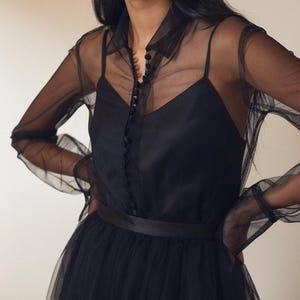 Puede incluir: Vestido negro en capas con una camisa transparente abotonada sobre un corpiño de tirantes finos. El vestido incluye una falda de tul y un cinturón de satén. Las mangas de la camisa transparente son largas y abullonadas. La modelo lleva pendientes plateados.