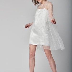 Puede incluir: Un minivestido blanco de satén con una superposición transparente adornada con pequeños puntos blancos. El vestido presenta un escote cuadrado y se combina con botines blancos. La modelo sonríe y parece estar en movimiento.