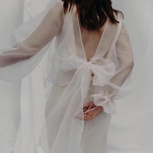 Puede incluir: Una blusa blanca transparente con mangas largas y fluidas y una espalda en V profunda. La blusa presenta un gran lazo en la cintura y detalles delicados en las muñecas. La prenda forma parte de una colección nupcial o de vestir formal.