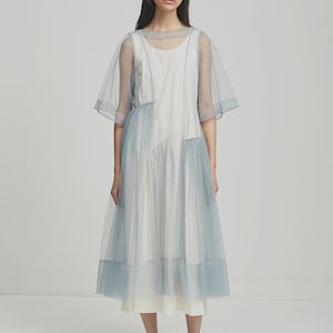 Puede incluir: Un vestido midi blanco y azul claro con mangas transparentes y una falda en capas. El vestido presenta un cuello redondo y un diseño asimétrico. La modelo lleva zapatos negros.