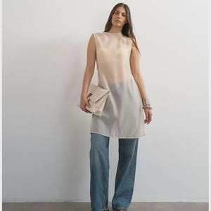 Puede incluir: Una túnica sin mangas, transparente y de color crema, usada sobre una prenda interior clara, combinada con jeans azules de corte holgado. El conjunto incluye un bolso de mano grande y plegado de color crema y una pulsera ancha de plata.