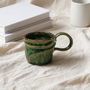 Puede incluir: Una pequeña taza de cerámica hecha a mano con un esmalte verde intenso brillante y una textura de superficie irregular y ondulada. La taza presenta un asa redondeada y un borde marrón oscuro, mostrando una forma orgánica y distintiva.