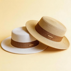 May include: Two straw hats with a brown band and a small gold-coloured tag. One hat is natural tan, and the other is white. The hats have a flat top and a wide brim. The background is a solid light yellow.