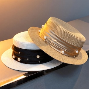 May include: Two straw hats with wide brims. One hat is white with a black ribbon and faux pearls. The other hat is tan with a gold ribbon and faux pearls. The hats are designed in a classic boater style.