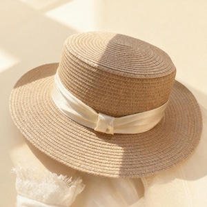 Op de afbeelding: Een beige strohoed met een brede rand en een cilindrische kroon. Een crèmekleurig lint is om de basis van de kroon gewikkeld en in een strik geknoopt. De hoed is ontworpen voor bescherming tegen de zon en mode.