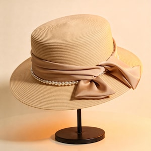 May include: A beige straw hat with a wide brim and a cylindrical crown. A satin ribbon in a matching beige color is wrapped around the crown, adorned with a string of faux pearls and a bow. The hat is displayed on a black stand.