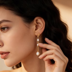 Op de afbeelding: Goudkleurige oorbellen met een bungelend ontwerp met witte parels. De oorbellen hebben een kleine hoepel die aan het oor wordt bevestigd, met een reeks parels en een rechthoekig goudkleurig element.