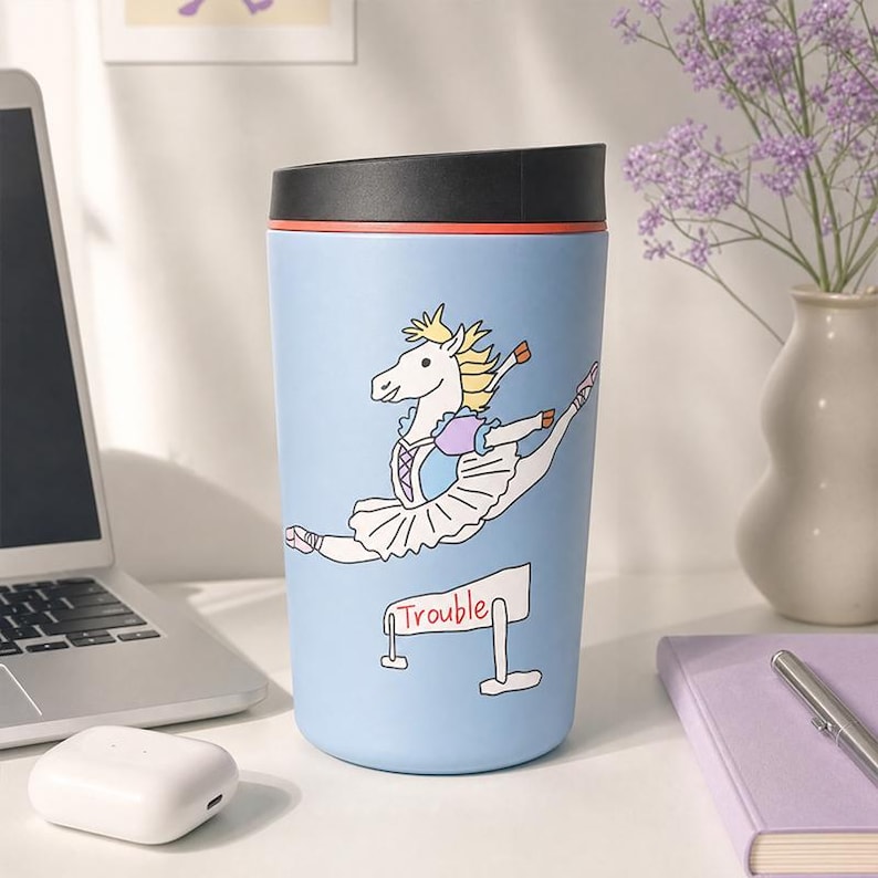 Pu&ograve; includere: Una tazza da viaggio azzurra con coperchio nero, caratterizzata da un'illustrazione a cartone animato di un cavallo bianco con tut&ugrave; da balletto e scarpette, che salta un ostacolo con la scritta "Trouble" in rosso sotto il cavallo.
