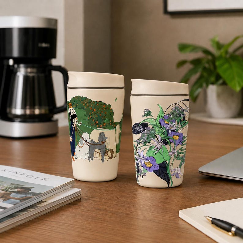 May include: Two off-white travel mugs with dark rims. The left mug features a person walking two dogs in a green park setting. The right mug displays a vibrant botanical illustration of purple flowers and green leaves against the cream background.