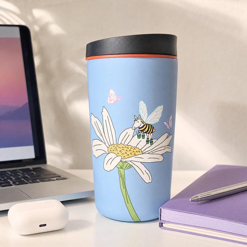 Pu&ograve; includere: Un bicchiere da viaggio azzurro con coperchio nero e bordo arancione. L'esterno presenta un'illustrazione grafica di una grande margherita bianca con stelo verde, una piccola ape con ali sui petali e due piccole farfalle di colore rosa.