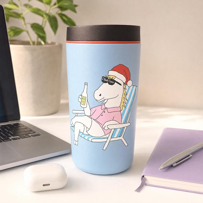 Pu&ograve; includere: Una tazza da viaggio azzurra con coperchio nero che presenta la grafica di un cavallo bianco con un cappello da Babbo Natale rosso e occhiali da sole neri con la scritta I Don't Care. Il cavallo &egrave; seduto su una sedia a sdraio a righe.