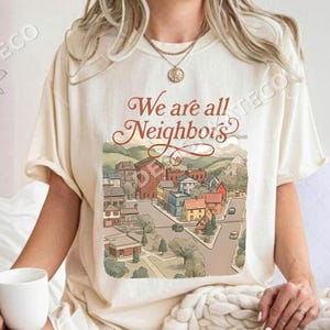 May include: Cream-coloured t-shirt with the text "We are all Neighbors" above a colourful illustration of a town with houses, trees, and roads. The shirt is made of a soft material.