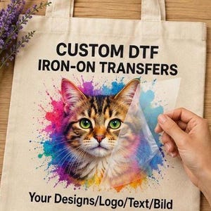 Könnte beinhalten: Eine beigefarbene Tragetasche aus Segeltuch mit einem bunten Aquarell-Spritzer-Design und einem getigerten Katzengesicht in der Mitte. Der Text CUSTOM DTF IRON-ON TRANSFERS steht über der Katze, Your Designs/Logo/Text/Bild steht unter der Katze.