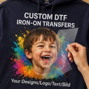 Könnte beinhalten: Ein dunkelblauer Kapuzenpullover mit einem individuellen DTF-Aufbügeltransfer. Die Grafik zeigt ein lachendes Kind, umgeben von einem bunten Farbspritzer-Effekt. Der Text lautet CUSTOM DTF IRON-ON TRANSFERS und Your Designs/Logo/Text/Bild.