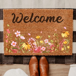 May include: A rectangular coir doormat with the word "Welcome" in black script. The mat features a watercolor floral design with pink, yellow, and purple flowers. The mat is placed on a black and white checkered rug.