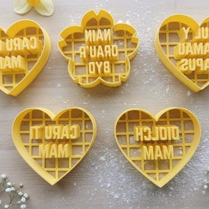 May include: Yellow plastic cookie cutters in heart and flower shapes. The cutters have text that reads "CARU CHOI MAM", "MAIN A UARD OYB", "Y JUZ UAMAM 2U9AH", "IT URAJ MAM", and "H3JOIO MAM".