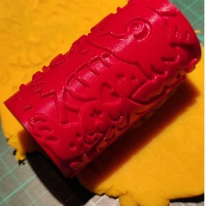 May include: A red patterned rolling pin on yellow playdough. The rolling pin features a detailed floral design. The playdough sits on a green cutting mat. The rolling pin is a craft tool.