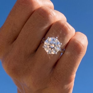 May include: A silver-toned ring featuring a large, round, brilliant-cut clear gemstone set in a six-prong setting. The stone displays multiple facets that reflect light, creating a bright, sparkling appearance against the clear blue sky background.