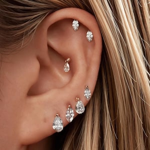 May include: A set of six gold-toned earrings featuring clear, pear-shaped and marquise-cut gemstones. The jewelry is arranged along the earlobe, conch, and helix, with each piece secured by a small metal post and backing in a delicate, minimalist style.