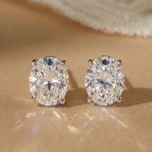 May include: A pair of oval-cut stud earrings featuring clear, faceted stones set in a metallic prong setting. The stones exhibit high brilliance and light reflection, creating a sparkling appearance with visible rainbow-colored light refractions.