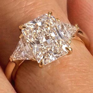 May include: A gold ring featuring a large rectangular radiant-cut center diamond held by four prongs, flanked by two smaller triangular-cut side diamonds. The metal band is polished yellow gold, showcasing a classic three-stone setting design.