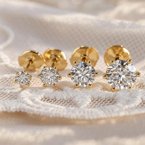 May include: Four gold stud earrings arranged in a row, each featuring a single round brilliant-cut clear gemstone held by a six-prong setting. The earrings are graduated in size from smallest to largest, showcasing polished gold posts and backs.