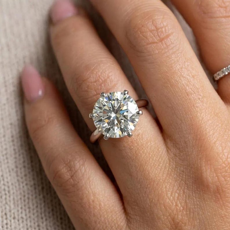 May include: A silver-toned ring featuring a large, round, brilliant-cut clear stone held in place by a six-prong setting. The band is smooth and polished, and a secondary, thinner band with small embedded stones is visible on the adjacent finger.