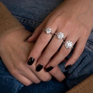 May include: Three silver-toned rings, each featuring a single large, round, brilliant-cut clear stone set in a six-prong setting. The rings are worn on the index, middle, and ring fingers, showcasing varying sizes of the central gemstone.