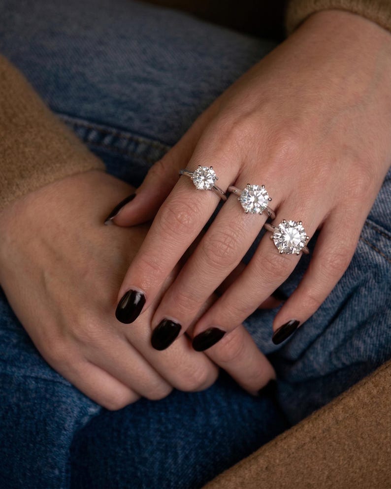 May include: Three silver-toned rings, each featuring a single large, round, brilliant-cut clear stone set in a six-prong setting. The rings are worn on the index, middle, and ring fingers, showcasing varying sizes of the central gemstone.