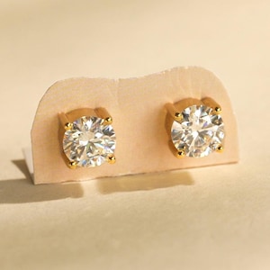 May include: A pair of stud earrings featuring round, clear, faceted stones set in four-prong gold metal settings. The earrings are mounted on a light beige display card, showcasing the brilliant cut of the stones and the polished gold finish.