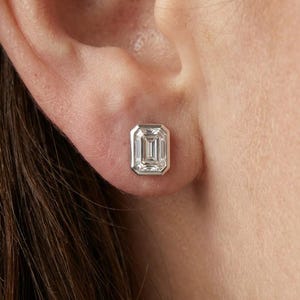May include: A close-up of a single emerald-cut diamond stud earring. The diamond is set in a silver or white gold bezel setting. The earring is shown on a person's ear.
