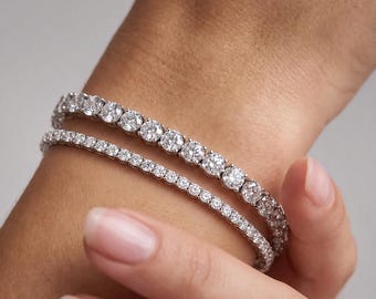 May include: Two silver-toned tennis bracelets featuring a continuous row of round, clear, faceted stones. The bracelets are layered together, with one featuring larger stones and the other featuring smaller stones, both set in a metallic band.