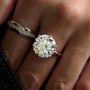 May include: A round brilliant cut solitaire ring with a six-prong setting on a polished metal band, accompanied by a secondary band featuring a channel-set row of small clear stones, both worn together on the finger to create a layered appearance.