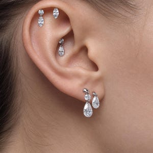 May include: A set of silver-toned metal earrings featuring clear, faceted stones in various shapes including marquise, round, and teardrop cuts. The jewelry is arranged across multiple piercings on the ear, including the lobe and the upper cartilage.