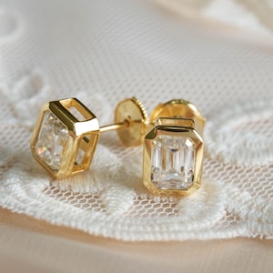 May include: A pair of gold stud earrings featuring emerald-cut clear stones set in a bezel setting. Each earring has a rectangular faceted center stone encased in a polished gold frame with a post back and a circular gold earring back closure.