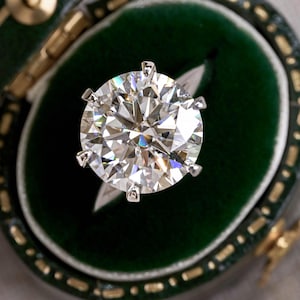 May include: A round brilliant cut diamond ring featuring a large, clear, faceted stone set in a six-prong silver-toned metal setting. The ring is displayed inside a dark green velvet-lined jewelry box with gold-toned trim along the edges.