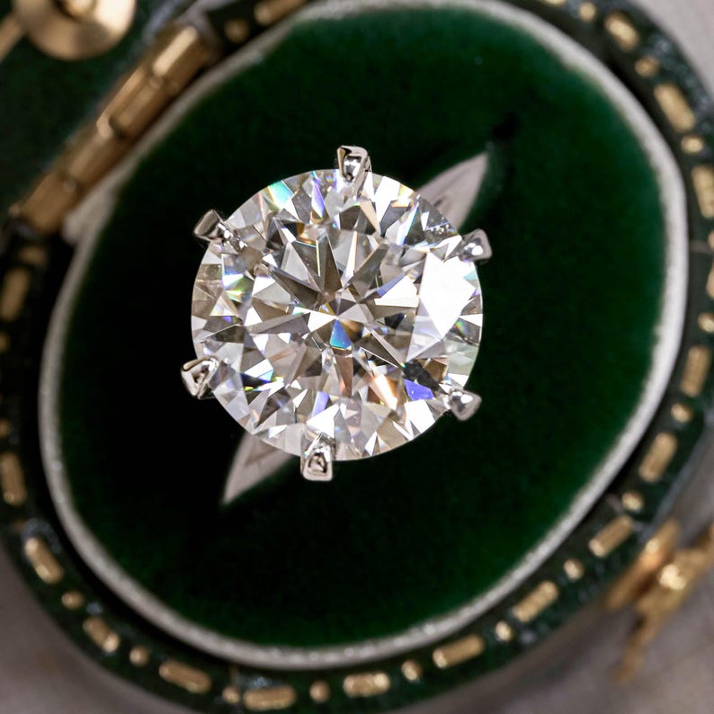 May include: A round brilliant cut diamond ring featuring a large, clear, faceted stone set in a six-prong silver-toned metal setting. The ring is displayed inside a dark green velvet-lined jewelry box with gold-toned trim along the edges.