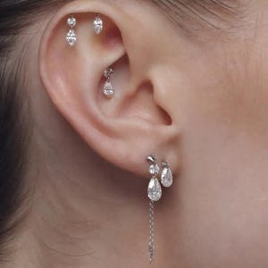 May include: A collection of silver-toned metal earrings featuring clear, faceted stones in various shapes including marquise, pear, and round cuts. The set includes multiple stud earrings placed along the ear cartilage and two dangling earrings.