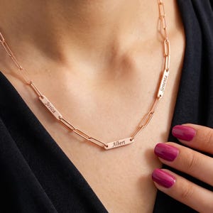May include: A rose gold-toned paperclip chain necklace featuring three rectangular metal bars engraved with the names Jesica, Albert, and Sementa. The necklace is constructed with elongated oval links and secures around the neck.